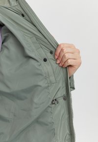 Lys grøn jakke indvendigt, der viser en glat foer, lynlås og trykknapper. Synlig hånd der holder jakken åben, med flere lommer.