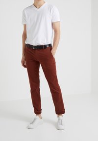 T-shirt bianco a V, chinos slim-fit color ruggine, cintura nera con fibbia argentata e sneakers bianche. Tessuto liscio, pantaloni arrotolati.