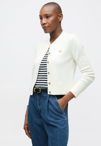 Mujer con cabello corto que lleva un cárdigan blanco abotonado sobre una camisa de rayas azul y blanca, jeans azules y un cinturón negro, de pie con una mano en el bolsillo.