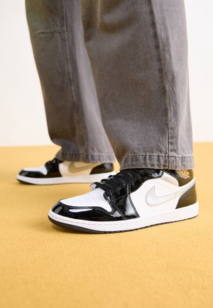 Personne portant des baskets Nike en cuir verni noir et blanc avec des lacets noirs et un pantalon gris, debout sur un sol jaune.