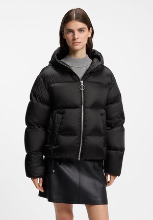 Junge Frau trägt eine schwarze Steppjacke mit Kapuze, grauen Pullover und schwarzen Lederrock vor einem einfarbigen hellen Hintergrund.