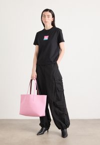 Black t-shirt with colorful logo, black cargo pants, and pink leather tote bag. Model wears pointed black heels. Smooth textures, modern design.