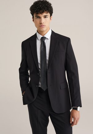 Zwarte colbert met bijpassende broek, wit overhemd en zwarte stropdas. Beschikt over een enkelvoudige sluiting, twee knopen en een subtiele textuur.