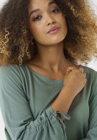 Green, long-sleeved shirt with a relaxed neckline and cinched sleeves, made of soft fabric, featuring a subtle texture and drawstring detail.