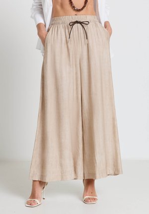 Beige Hosen mit weitem Bein, elastischem Bund und Kordelzug, kombiniert mit weißen offenen Absätzen und einem weißen Hemd, Hände in den Taschen.