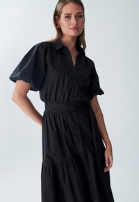 Abito nero a camicia con colletto, maniche corte a sbuffo, chiusura frontale con bottoni e cintura annodata, caratterizzato da un dettaglio a balze nella gonna.