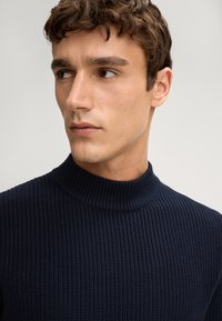 Marineblaue, gerippte Sweater mit Stehkragen aus strukturiertem Material, eng anliegendem Design und glattem Halsausschnitt. Fokus auf dem Oberkörper und dem Halsbereich.