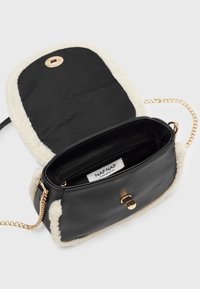 Svart väska med fluffig vit kant, utrustad med ett gult lås, kedjeförsedd axelrem och svart inredning med en sidoficka med dragkedja och etikett.