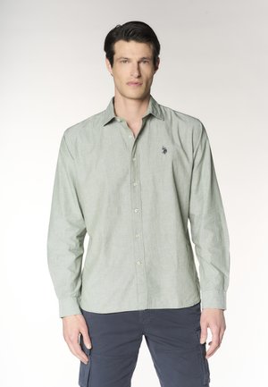 Uomo che indossa una camicia verde chiaro con bottoni e pantaloni cargo scuri, in piedi davanti a uno sfondo chiaro uniforme.