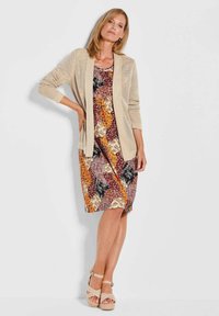 Beige gestrickte Strickjacke, die über einem mehrfarbigen, gemusterten Kleid mit floralem und geometrischem Design getragen wird. Das Modell trägt beige Plateausandalen.