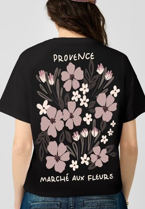 Person trägt ein schwarzes Shirt mit pinkem und weißem Blumenmuster und dem Text "Provence Marché aux Fleurs" auf dem Rücken.