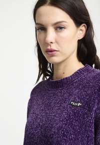 Lilla strikket sweater med en tekstureret overflade og en rund halsudskæring. Har et lille sort og hvidt "Volcom" logo på brystet.