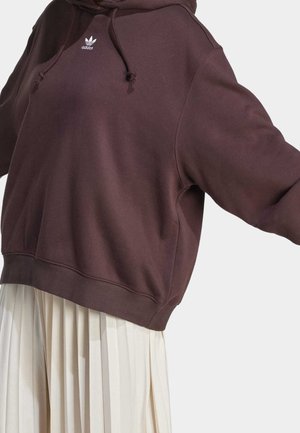 Sudadera con capucha Adidas de color marrón oscuro con logo en el pecho, llevada sobre una falda blanca plisada, brazos de la persona extendidos hacia afuera.