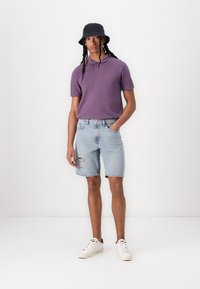 Purple polo shirt with two buttons, light blue ripped denim shorts, white sneakers, and a black bucket hat, posing against a white background.