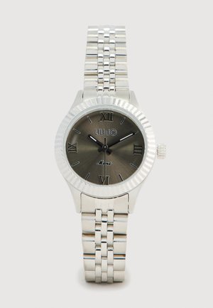 Srebrny zegarek z żłobkowaną lunetą, ciemnoszarym cyferblatem, cyframi rzymskimi i metalową bransoletą. Wyróżniające się logo marki na godzinie 12.