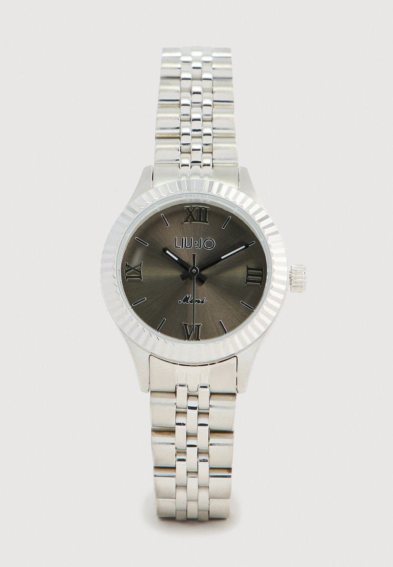 Orologio argento con lunetta scanalata, quadrante grigio scuro, numeri romani e bracciale a maglie metalliche. Logo del marchio ben visibile alle 12.