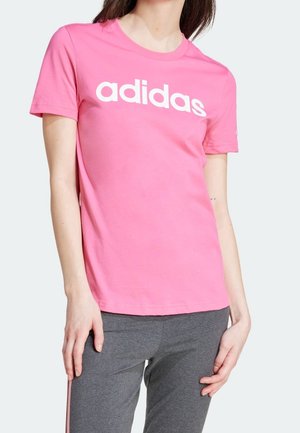 Femme portant un T-shirt Adidas rose avec un logo sur la poitrine et un pantalon de sport gris, debout, les mains détendues le long du corps.