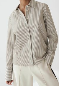 Personne portant une chemise à manches longues rayée verticalement crème et noire avec de larges poignets et un pantalon blanc plissé, debout avec une main dans la poche.