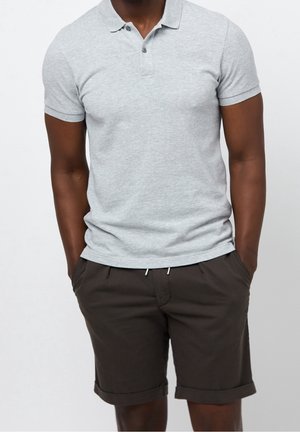 Uomo che indossa una polo grigio chiaro a maniche corte e pantaloncini marrone scuro con orlo arrotolato, con le mani nelle tasche, su uno sfondo semplice.