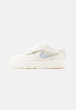 Valge madala säärega toss valgete paelte ja poolläbipaistva halli Nike Swoosh logoga külgedel, ühelisel heledal taustal.