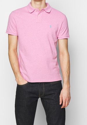 Homme portant un polo rose à manches courtes avec un petit logo bleu brodé, associé à un jean bleu foncé, debout devant un fond uni.