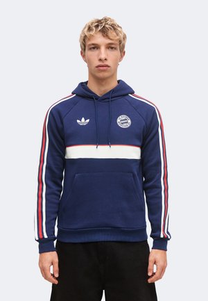 Giovane uomo con capelli biondi corti che indossa una felpa con cappuccio blu navy con strisce rosse e bianche e loghi FC Bayern Monaco e Adidas.
