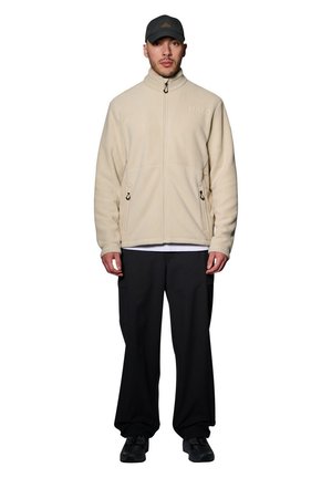 Beige fleece jack met hoge kraag, ritssluiting en zijzakken. Draagt over een wit shirt en gecombineerd met een zwarte broek en een grijze pet.