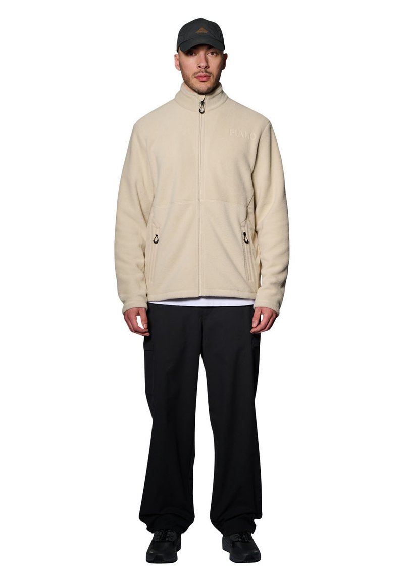 Beige fleecetröja med hög krage, dragkedja och sidofickor. Bärs över en vit skjorta och kombineras med svarta byxor och en grå keps.