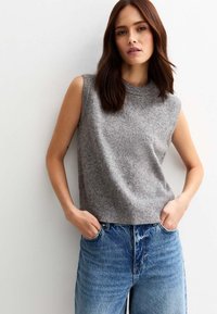 Mouwloze grijze brei-top met een hoge halslijn, gecombineerd met lichtblauwe high-waisted jeans met vervaagde patronen en grote zakken.