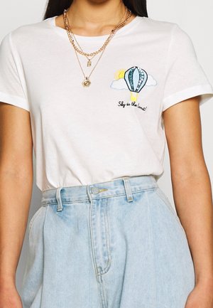 Kvinde iført hvid T-shirt med luftballon og teksten "Sky is the limit", kombineret med lyseblå højtaljede cowboybukser.