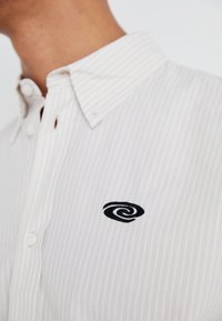 Camicia bianca con bottoni, presenta un piccolo logo a spirale nera, con delle sottili strisce verticali grigio chiaro e un colletto classico dal texture morbida.
