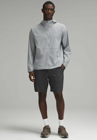 Giacca zip-up grigia chiara con cappuccio, abbinata a pantaloni corti scuri. Il tessuto sembra morbido e tecnico, ideale per attività all'aperto.