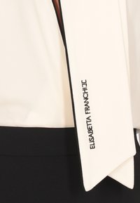 Blusa bianca con un nastro nero come accento e nome del marchio ricamato, caratterizzata da una texture liscia e un design pulito.