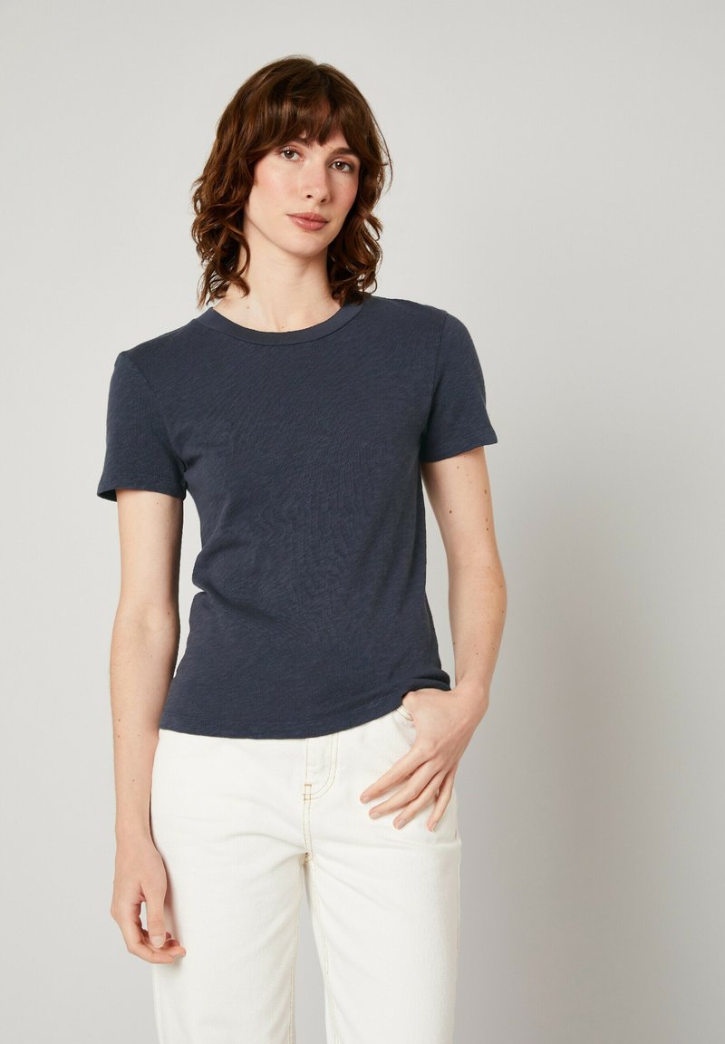 Tumši zila īsām piedurknēm t-kreklu ar apaļu kakla izgriezumu, izgatavots no mīksta, teksturēta auduma. Jānēs kopā ar gaišiem, pieguļošiem džinsiem.