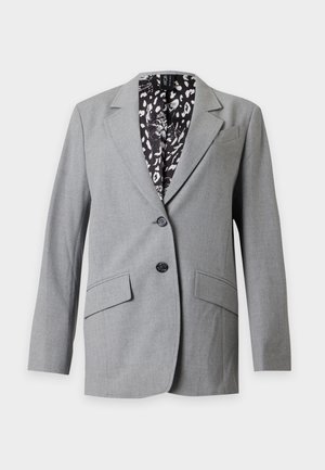 Femme aux cheveux bouclés portant un blazer gris, une chemise blanche et noire à motifs, un pantalon en cuir noir et des bottes noires pointues, debout devant un fond uni.