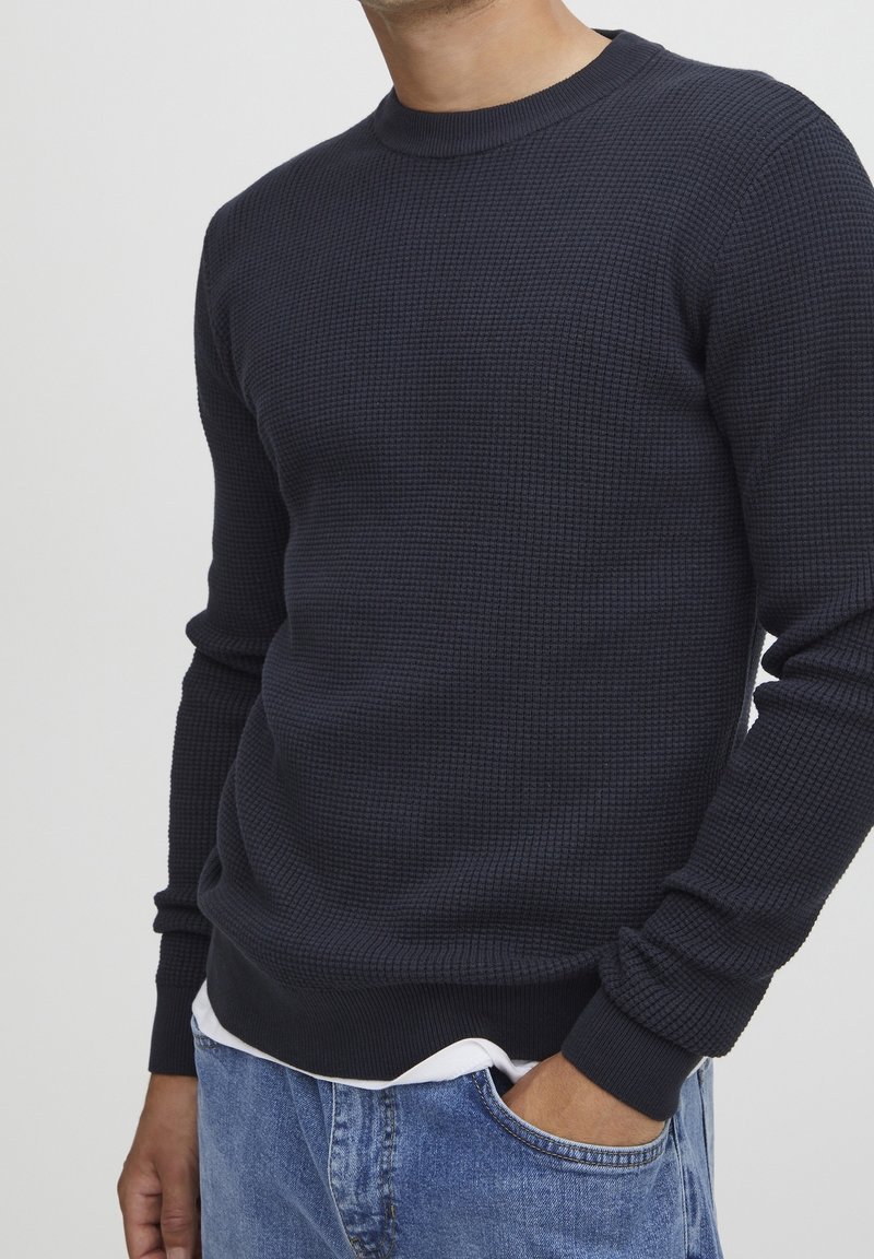 Homme portant un pull en maille texturée bleu marine foncé par-dessus une chemise blanche, avec une main dans la poche d'un jean bleu clair.