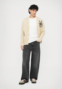 Cardigan en tricot beige avec une grande broderie florale, porté sur une chemise blanche, associé à un jean noir ample et des chaussures multicolores.