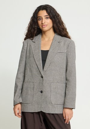 Femme aux longs cheveux bouclés portant un haut noir, un blazer oversize à motif pied-de-poule et un pantalon ample foncé sur un fond uni.