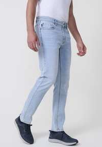 Jeans de mezclilla azul claro con un corte ajustado, que cuentan con un cierre de cremallera, cinco bolsillos y detalles de costura en contraste. Combinados con zapatillas oscuras.