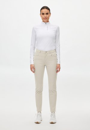 Kvinde stående mod fronten iført en hvid langærmet lynlåstrøje, beige smalle bukser og hvide sneakers på en hvid baggrund.