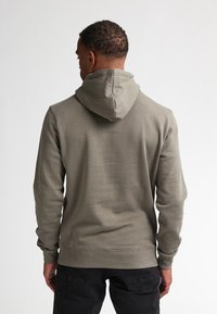 Sudadera verde oliva con capucha ajustable, con un borde ribeteado en el dobladillo y los puños, hecha de un tejido suave y texturizado. Se lleva debajo unos jeans negros.