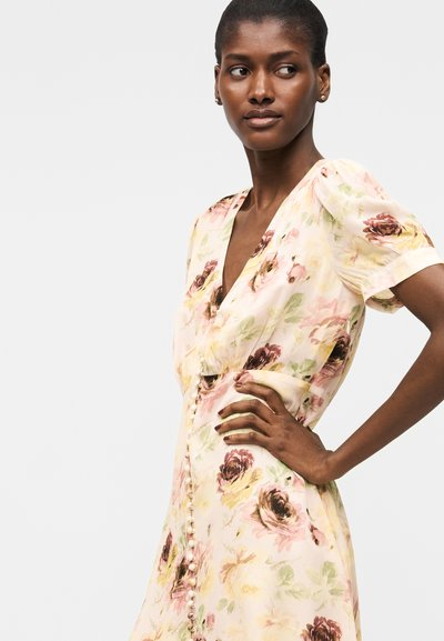 Γυναίκα με σκούρο δέρμα και κοντά μαλλιά που φοράει ελαφρύ φλοράλ φόρεμα με φουσκωτά μανίκια και κουμπιά μπροστά, στέκεται με το χέρι στη μέση.