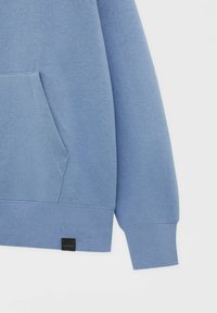 Manche de sweat-shirt bleue et poche kangourou avec une étiquette noire du logo Pull&Bear sur l'ourlet inférieur sur fond blanc.