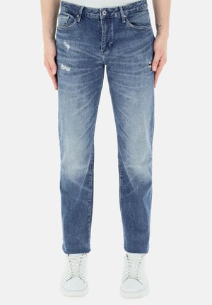 Jeans in denim blu con un taglio slim, caratterizzati da aree scolorite e un leggero sbiadimento. Indossati con sneakers bianche, mostrano uno stile casual.