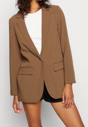 Frau trägt braunen Blazer mit einem Knopf über weißem Oberteil und schwarzen Shorts, steht vor schlichtem weißem Hintergrund.