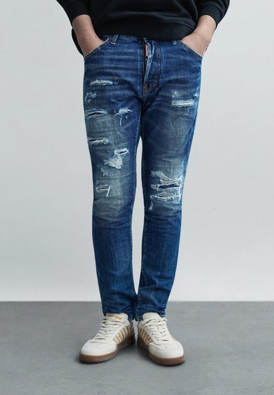 Mellanblå denimjeans med slitna partier, smal passform, femficksmodell, slitdetaljer, och kombinerade med beige sneakers med guldrand.