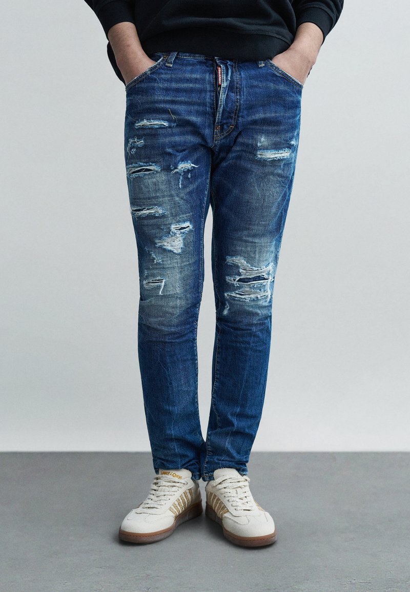 Φθαρμένο μπλε τζιν με ξεβαμμένες τρύπες, στενή εφαρμογή, σχέδιο με πέντε τσέπες, φθαρμένες λεπτομέρειες, συνδυασμένο με μπεζ αθλητικά παπούτσια με χρυσές ρίγες.