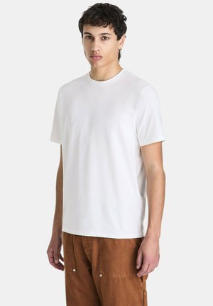 Jonge man met kort krullend haar die een effen wit T-shirt met korte mouwen en een bruine broek draagt tegen een effen achtergrond.