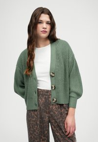 Groene gebreide cardigan met een relaxte pasvorm, voorzien van een diepe V-hals, drie grote knopen en ribbelmanchetten, gecombineerd met een patroonbroek.