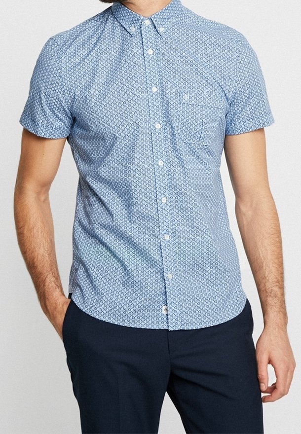 Homme portant une chemise bleue à manches courtes à motifs avec une poche poitrine et un pantalon sombre, debout les mains dans les poches.